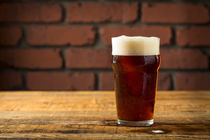 A pint glass of dark beer on a table next to brick wall.
