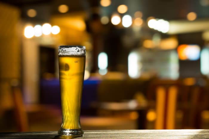 A tall glass of light colored beer in a bar or pub setting.