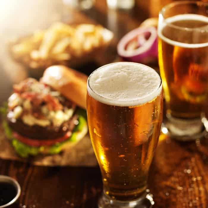 Two glasses of light-colored beer with a burger and fries in the background.